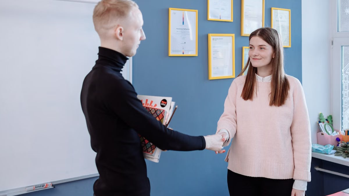 Man and woman exchanging handshake in office environment with certificates on wall.